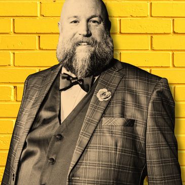 Bearded man in a checkered suit stands against a yellow brick wall.