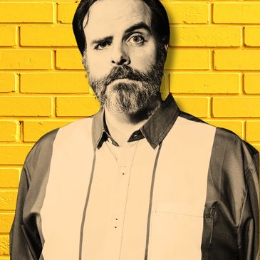 Man with a beard and raised eyebrow against a yellow brick wall.