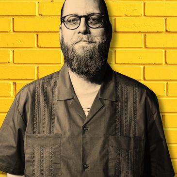 Bearded man with glasses in front of a bright yellow brick wall.