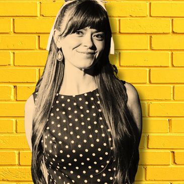 Woman in a polka dot dress smiling against a yellow brick wall.