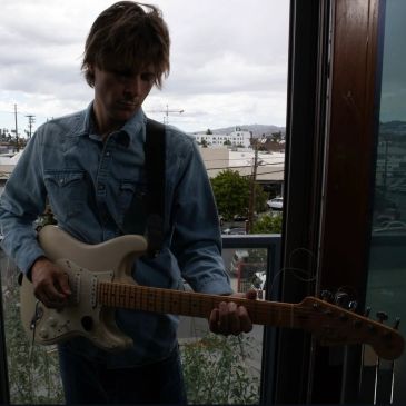 Man playing electric guitar near a window in a denim shirt.