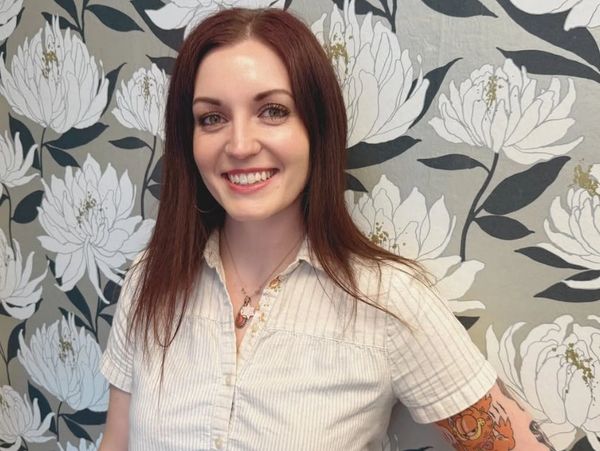 Smiling woman with red hair and tattoo, standing in front of floral wallpaper.