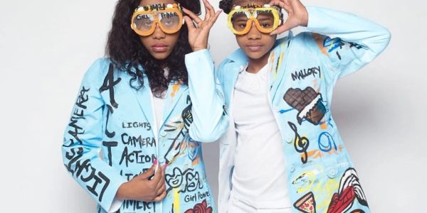 Two women wearing colorful graffiti-style jackets and oversized yellow glasses.