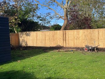 Garden with green grass, wooden fence, wheelbarrow, and tools on a sunny day.