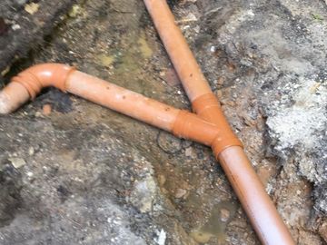 Exposed orange drainage pipes in muddy ground near a brick wall.