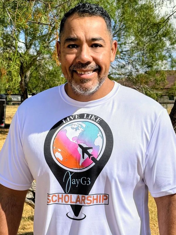 Smiling man wearing a 'Live Like JayG3 Scholarship' t-shirt outdoors.