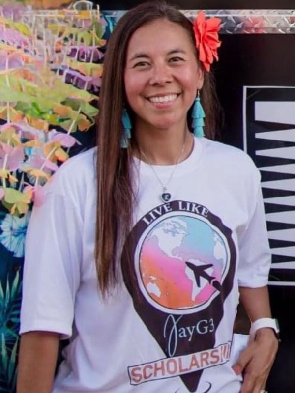 Smiling woman with flower in hair wearing a white JayG3 Scholars T-shirt.
