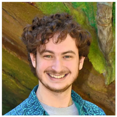 Young man with curly hair smiling outdoors in front of a mossy rock.