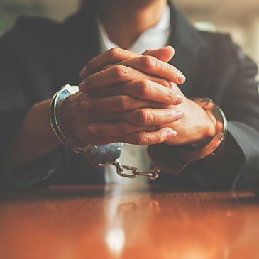 Man in black suit with hands cuffed in front of him at table