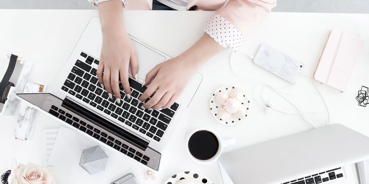 Hands typing on a laptop with coffee and snacks nearby on a white desk.