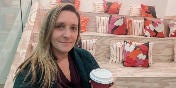 Woman holding a coffee cup, sitting on wooden steps with colorful cushions.
