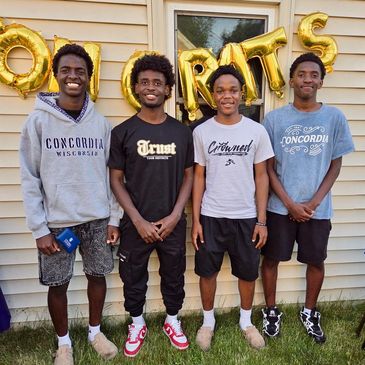 Four young men smiling in front of a congratulatory balloon backdrop outdoors.