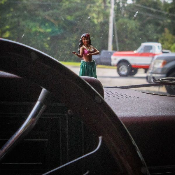 Hula girl dashboard doll inside a car with trucks outside.