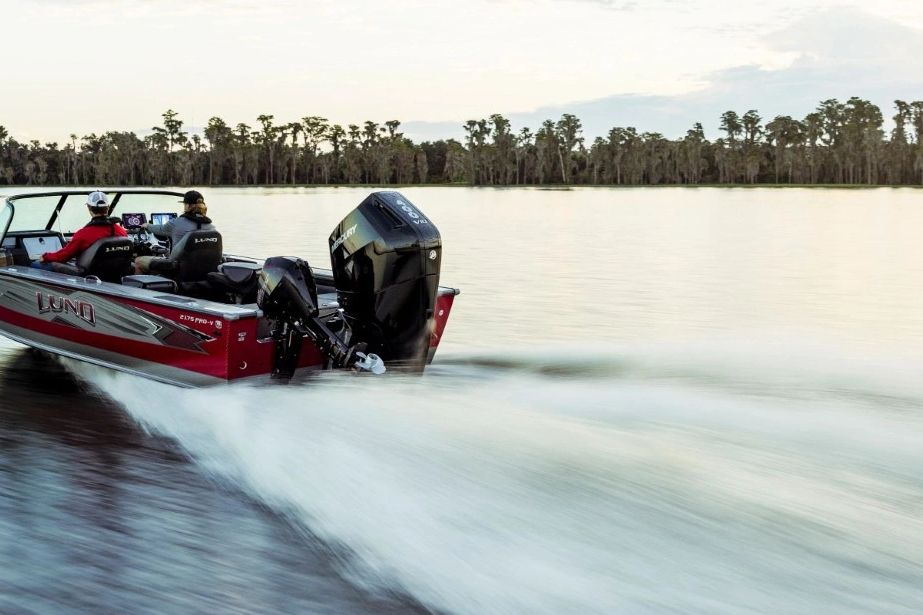 Two people ride a red Lund motorboat across calm water at sunset.