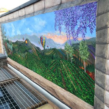 Outdoor block wall painted with DeLuz vineyard scene, purple tree, sunrise, hot air balloons.