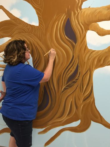Jill Roberts artist painting a wall mural of large tree on blue sky at Rancho Springs Medical Center