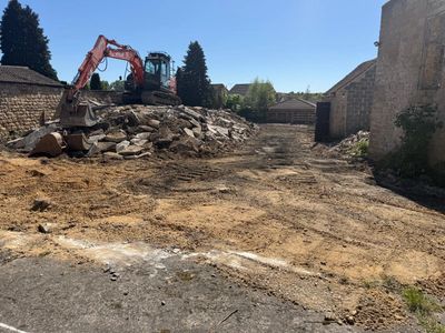 Excavator clearing rubble in a sunny outdoor area surrounded by stone buildings.