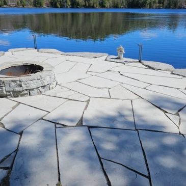Stone fire pit on a patio overlooking a calm lake with trees.