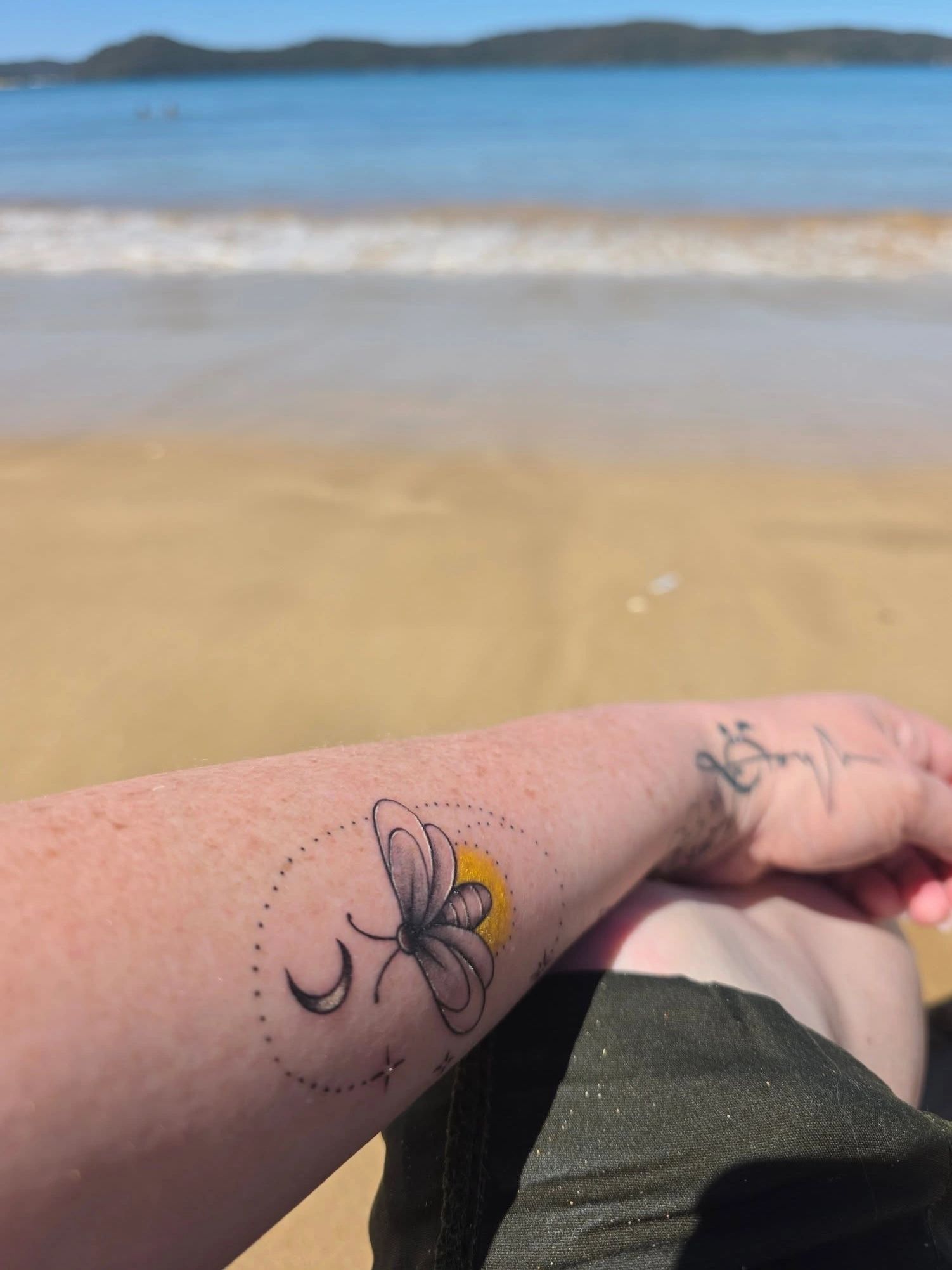 Arm tattoo of a bee with crescent moon and yellow background at the beach.