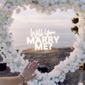 A romantic marriage proposal with a heart-shaped floral frame and a hand showing an engagement ring.