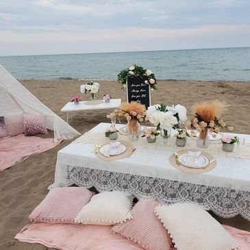 Elegant beach picnic setup with lace tablecloth and pastel cushions.