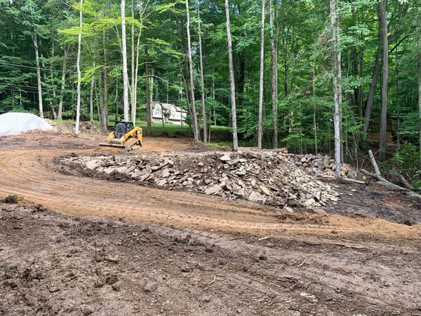 Construction site clearing brush, debris, stumps, and a pile of rocks and a skid-steer loader.