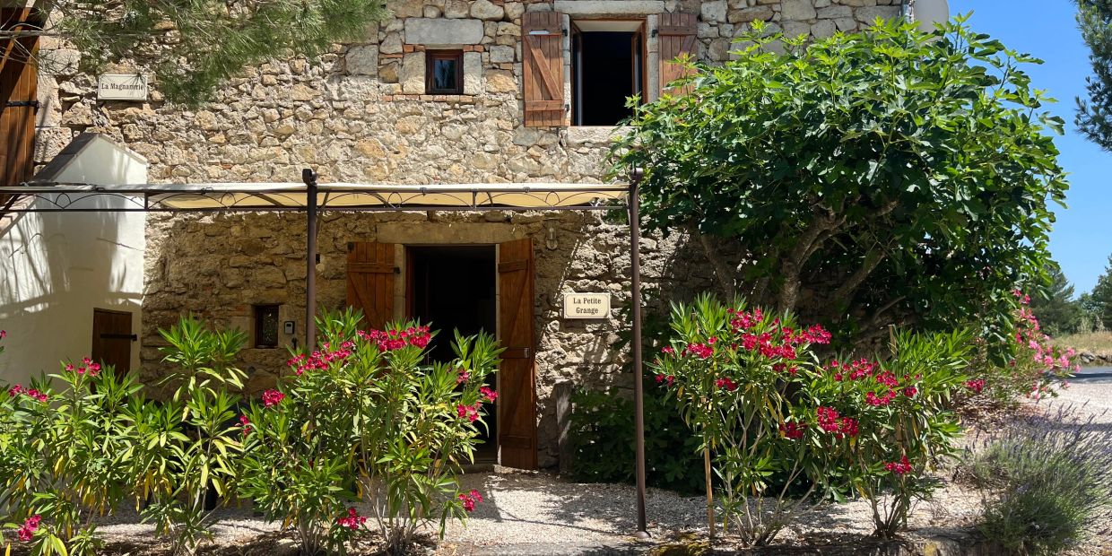 Stone house with wooden shutters surrounded by flowering bushes and greenery.