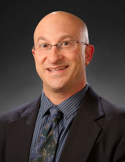 Smiling bald man in glasses, suit, and patterned tie against a dark background.
