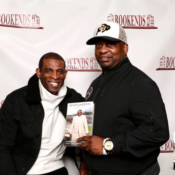 Two men posing together, one holding a book by Deion Sanders.