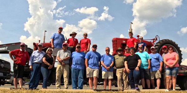 Group of people posing outdoors with tractors under a partly cloudy sky.