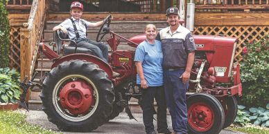 Three generations on a vintage tractor outside a wooden house.