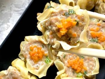 Chopsticks holding a piece of shumai dumpling topped with orange garnish.