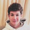 A young man with curly hair smiles warmly at the camera indoors.