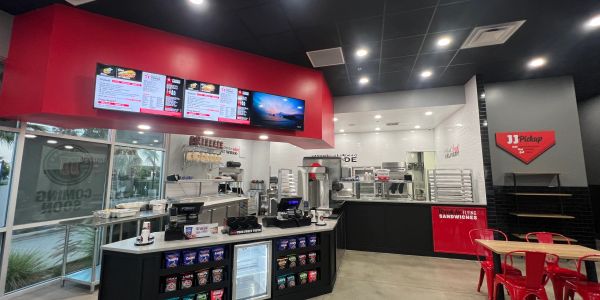 Modern sandwich shop counter with menu screens and red chairs.