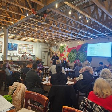 A community meeting with a speaker and an agenda displayed on a screen in a lively venue.