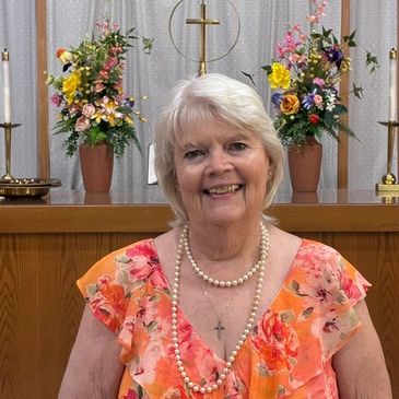 Smiling elderly woman in floral dress with pearl necklace in church setting.