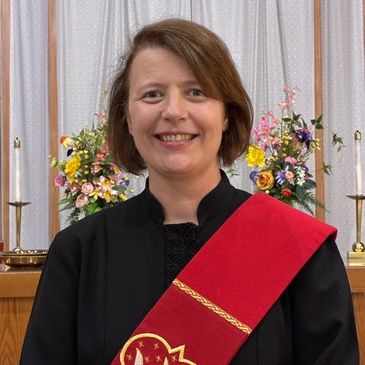 A smiling woman in religious attire with a red stole stands in a church.