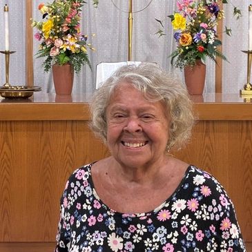 Smiling elderly woman in floral dress inside a church with flower arrangements.