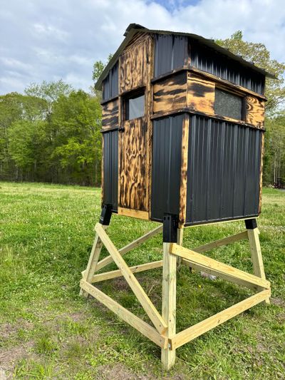 A wooden hunting blind elevated on a sturdy wooden frame in a grassy field.