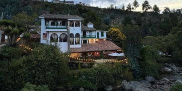 A cozy, illuminated house nestled among trees at dusk near rocky terrain.