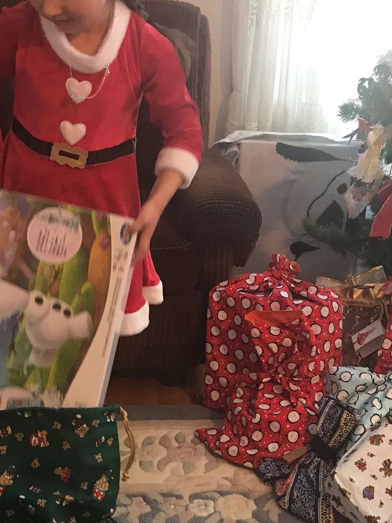 Child in a Santa dress opening a Christmas gift near a decorated tree.