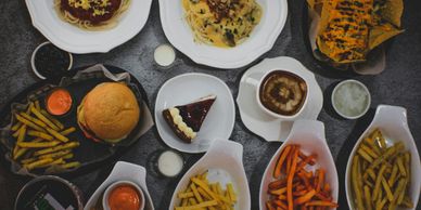 A variety of dishes including pasta, burger, fries, cake, and coffee on a dark table.