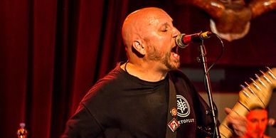 Bald man passionately singing and playing guitar on stage with red curtain backdrop.