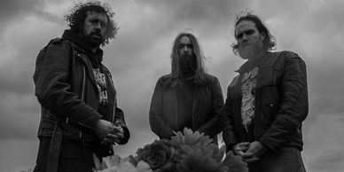 Three men in dark clothing pose under a cloudy sky in a black and white photo.