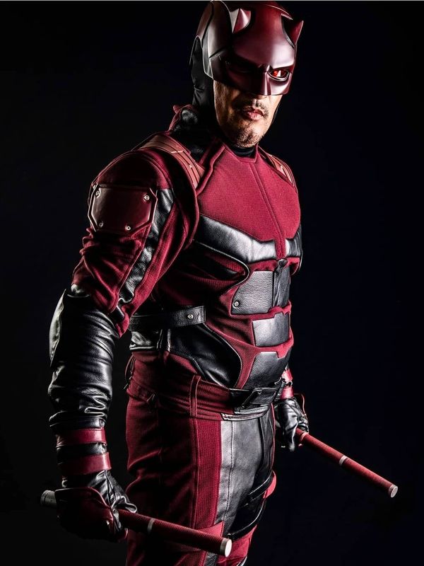 A man dressed as Daredevil holding two batons against a dark background.