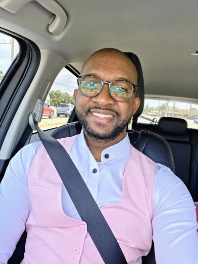 Man wearing glasses and a pink vest smiles in a car.