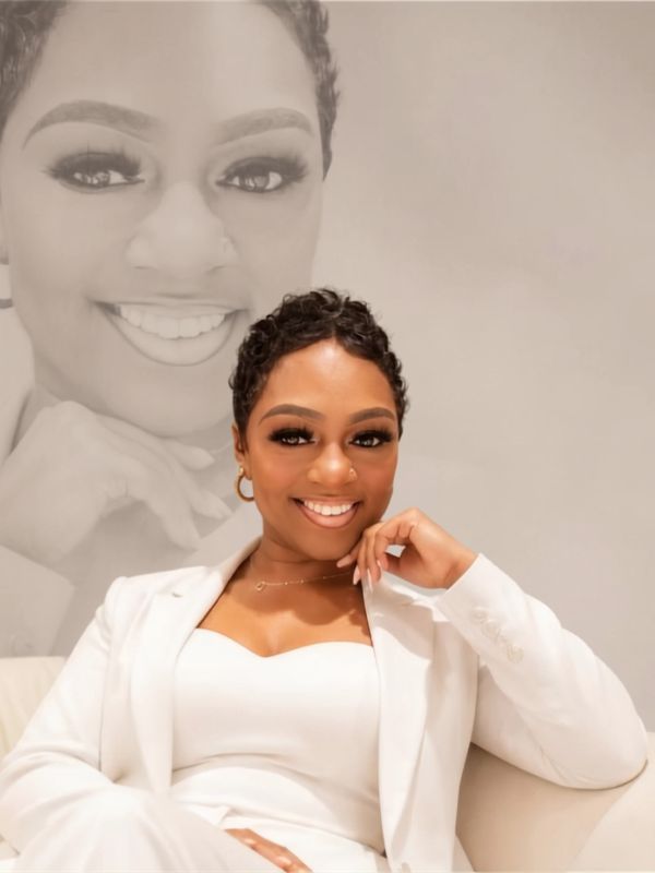 Confident woman in white suit smiling with a large portrait in the background.