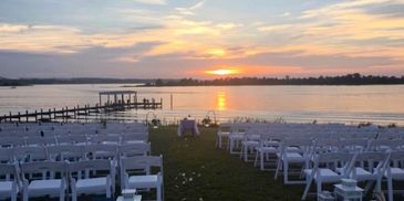 Outdoor wedding setup by the water at sunset with white chairs and lanterns.