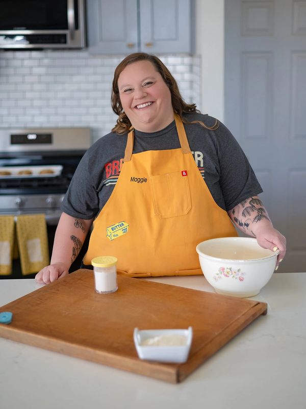 Smiling woman wearing a yellow apron in a kitchen with baking ingredients.
