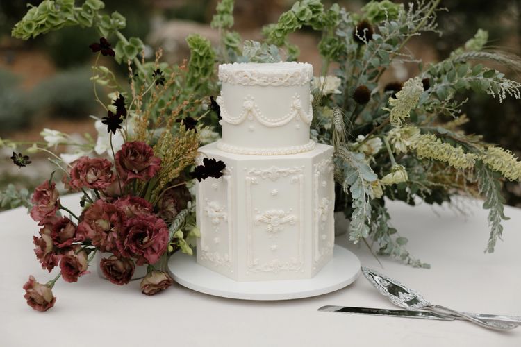 Two-tier vintage white wedding cake with ornate floral designs and surrounding flowers.